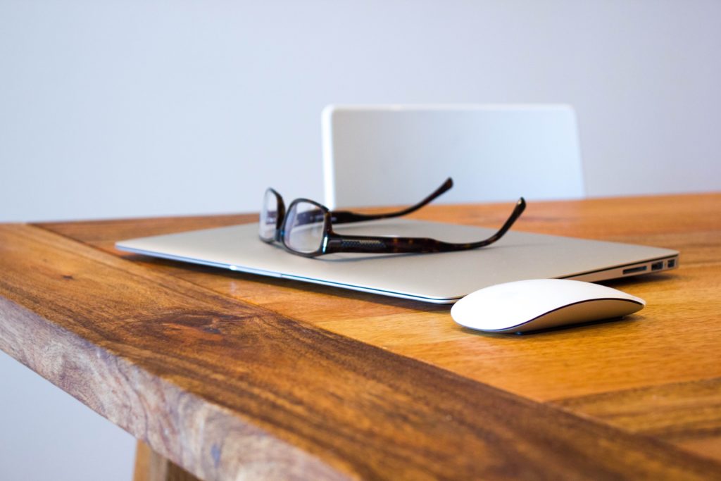 Laptop, wireless mouse, and eyeglasses on top of a wooden table.