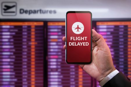 Businessman using Mobile Phone, where "flight delayed" text is on the screen, in front of Departures Board